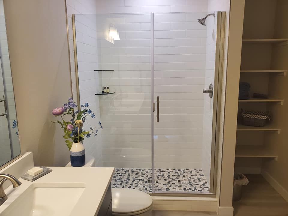 Bathroom with white tiled shower, glass door, and a storage shelf. Blue and white vase with flowers on countertop.