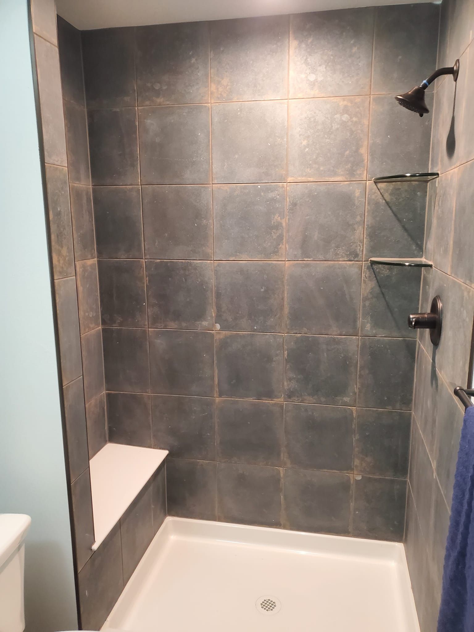 Shower with dark gray tile, white shower pan, seat, and shelves, and bronze fixtures.