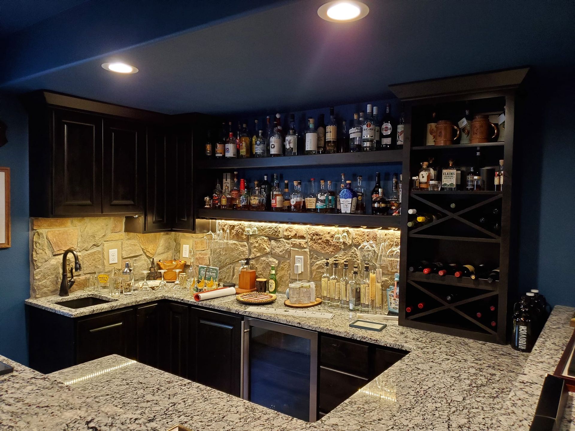 Dark wood home bar with granite countertop, stocked shelves against a blue wall.