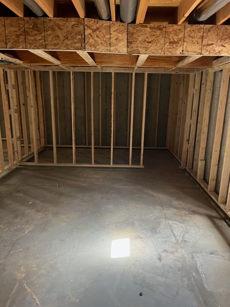 Unfinished basement room framed with wood studs, concrete floor, exposed joists overhead, and utility pipes.