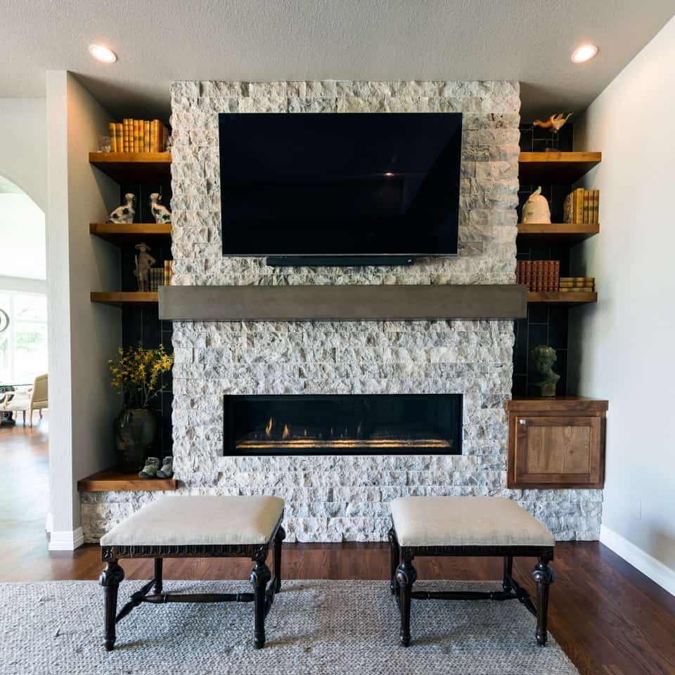 Fireplace with stone surround, TV, shelves with decor, and two upholstered ottomans in a modern living room.