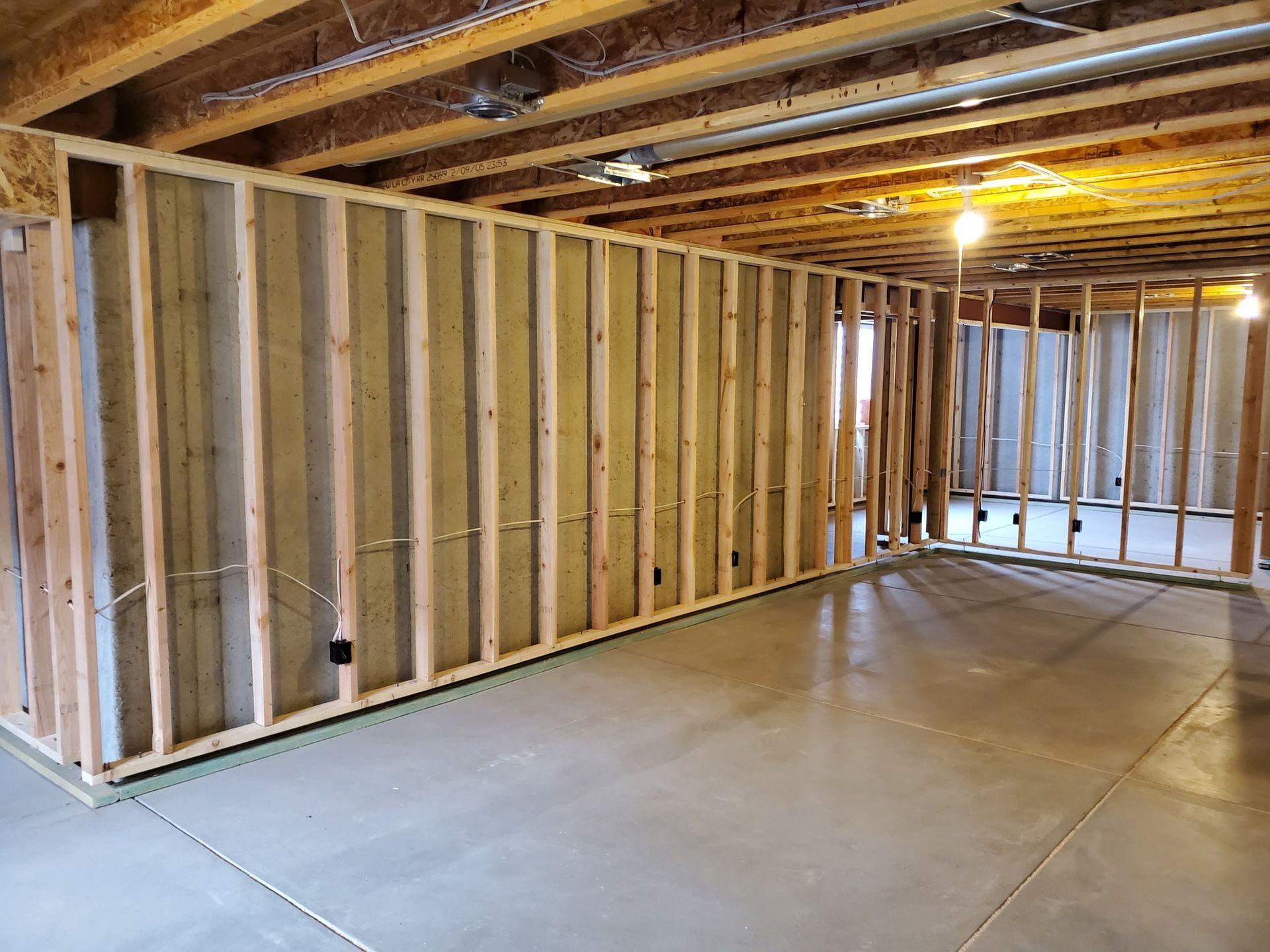 Interior of a room with unfinished wooden framing and a concrete floor. Overhead lights are visible.