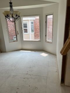 Empty room with bay window. Chandelier hangs above unfinished floor, overlooking brick building.