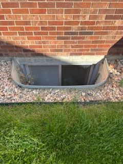 Window well in a brick wall with a screen and gravel, next to a grassy lawn.