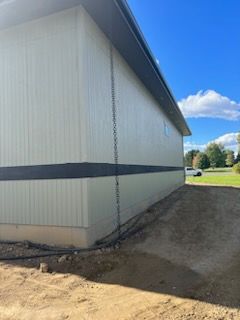 Side of a beige commercial building with a black horizontal stripe and a chain downspout; dirt and blue sky.