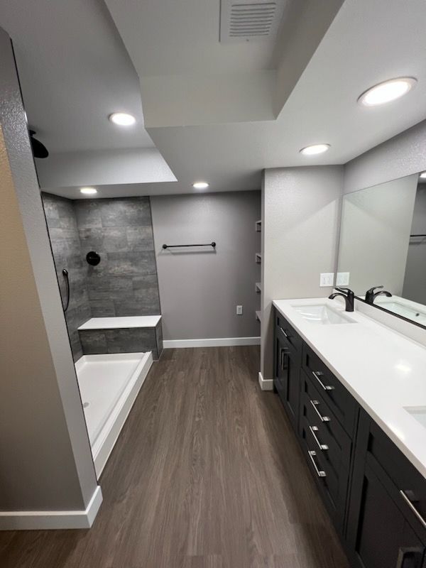 Modern bathroom with dark gray vanity, white countertop, tiled shower, and wood-look flooring.