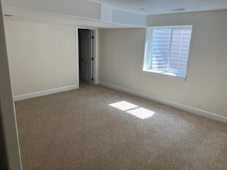Empty room with beige carpet and off-white walls. Window lets in sunlight. Doorway visible in the back.
