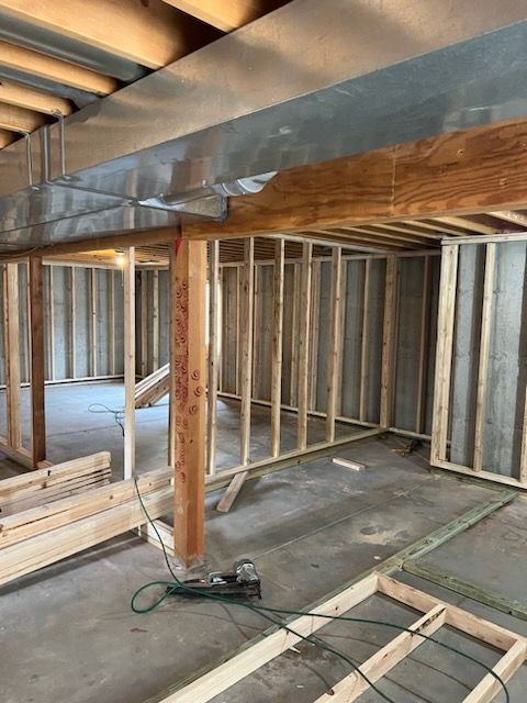 Basement construction: wooden framing, concrete floor, metal ductwork overhead. Tools and lumber scattered about.