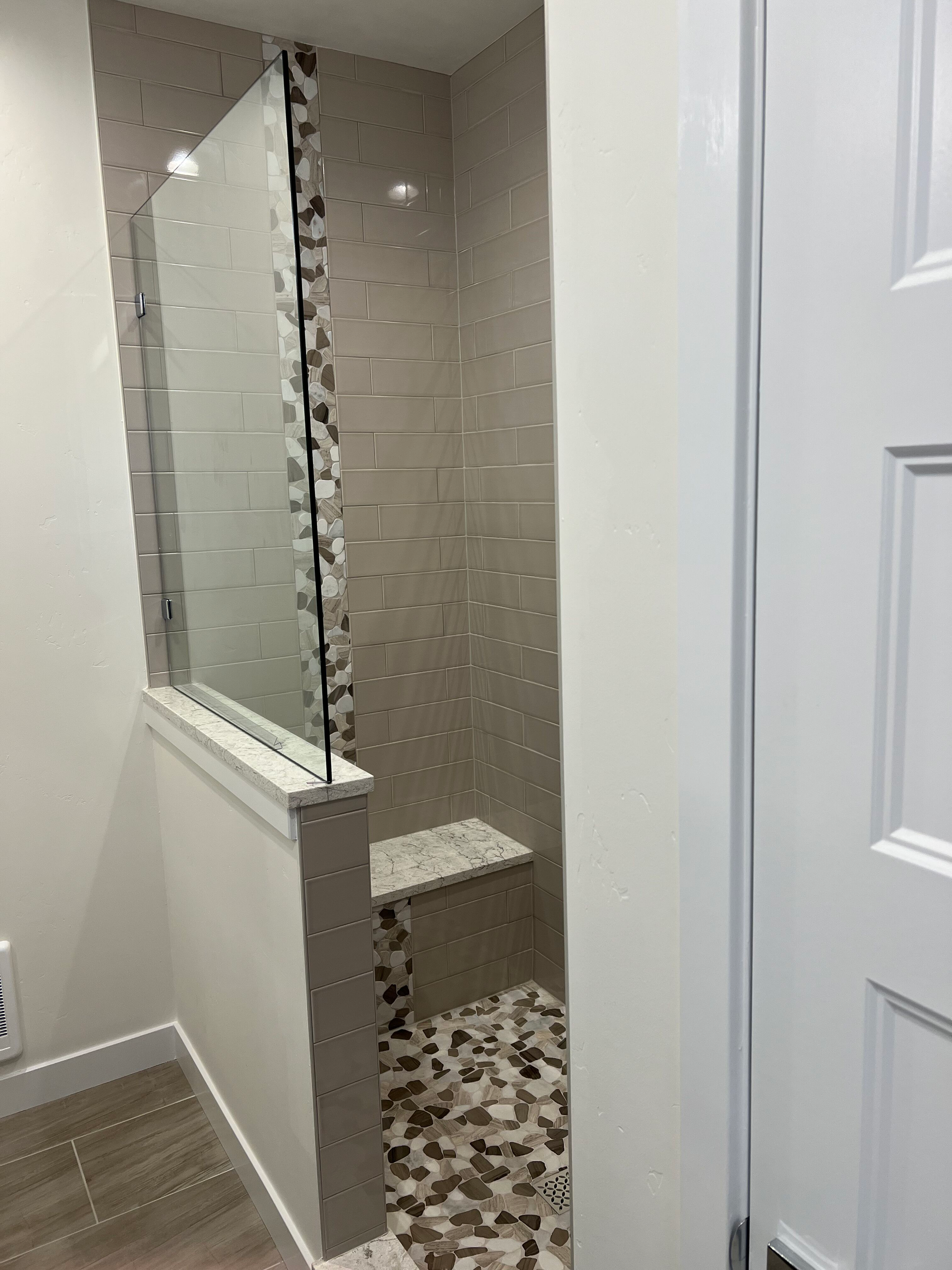 A modern shower with glass panel, pebble floor, tile walls, and built-in bench in a bathroom.