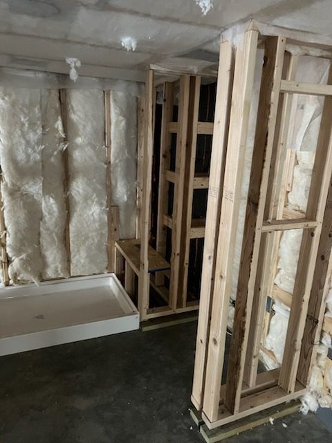Bathroom remodel: unfinished walls with insulation and new shower base. Wooden framing. Concrete floor, overhead lighting.