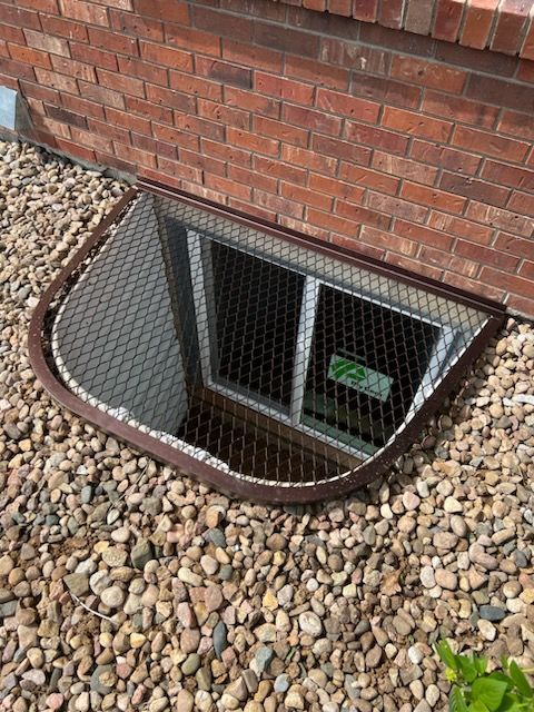 Brown metal window well with wire mesh cover against a brick wall, surrounded by gravel.