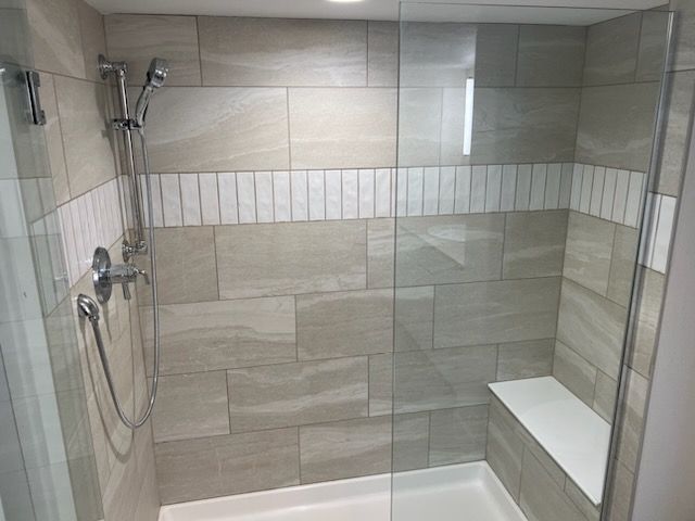 Shower with beige stone tiles, a white stripe, and glass enclosure. A built-in bench is visible.