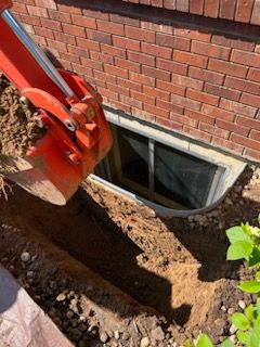 An orange excavator digging trench near a brick wall with a basement window.