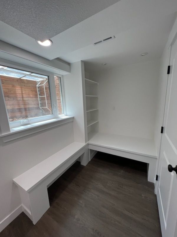 White built-in bench seat with shelves, by a window in a bright room, dark flooring.