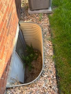 Metal window well next to a brick wall, filled with gravel, grass on right.