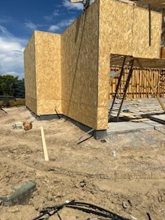 Building under construction; exterior walls of OSB wood, dirt ground, blue sky in background.