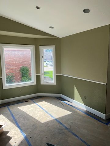Room interior, sage green walls, white trim, two windows, bare floor with tape.