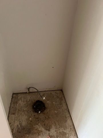 Small, empty room with exposed concrete floor and toilet drain. A pipe is connected. White walls.
