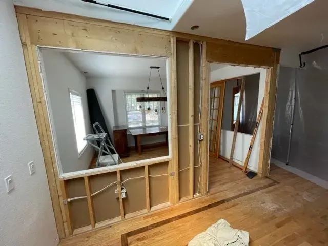 Interior view of a room under renovation. Wall removed, revealing a dining room; exposed studs and wiring.