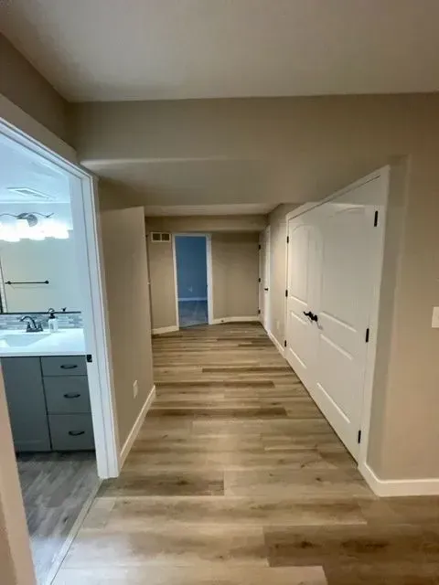 Hallway with light brown wood flooring, beige walls, and white doors. Open doorway to bathroom on the left.