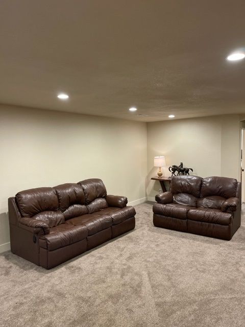Brown leather sofa and loveseat on a carpeted floor in a room with beige walls. A lamp and side table are in the background.