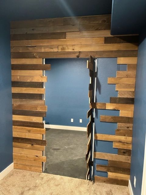 Wooden wall with a doorway opening into a blue-walled room. Floor is carpeted, the doorway is wooden.