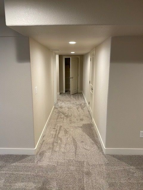 A long, narrow hallway in a basement with light gray carpet, white walls, and recessed lighting.