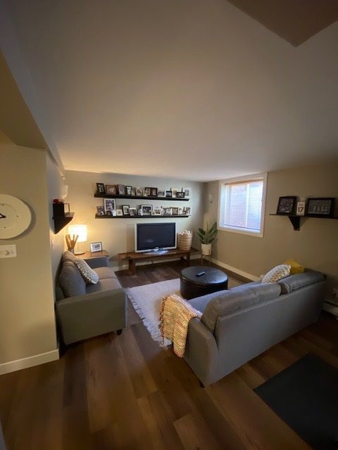 Living room with gray couches, dark wood floors, TV, shelves with photos, and a window.