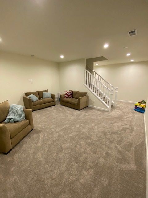 Spacious room with tan carpet, three brown couches, and a staircase leading up. Beige walls.