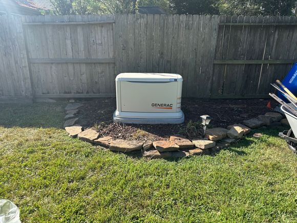 A beige home generator on a gravel bed, bordered by stones, in a backyard near a wooden fence.