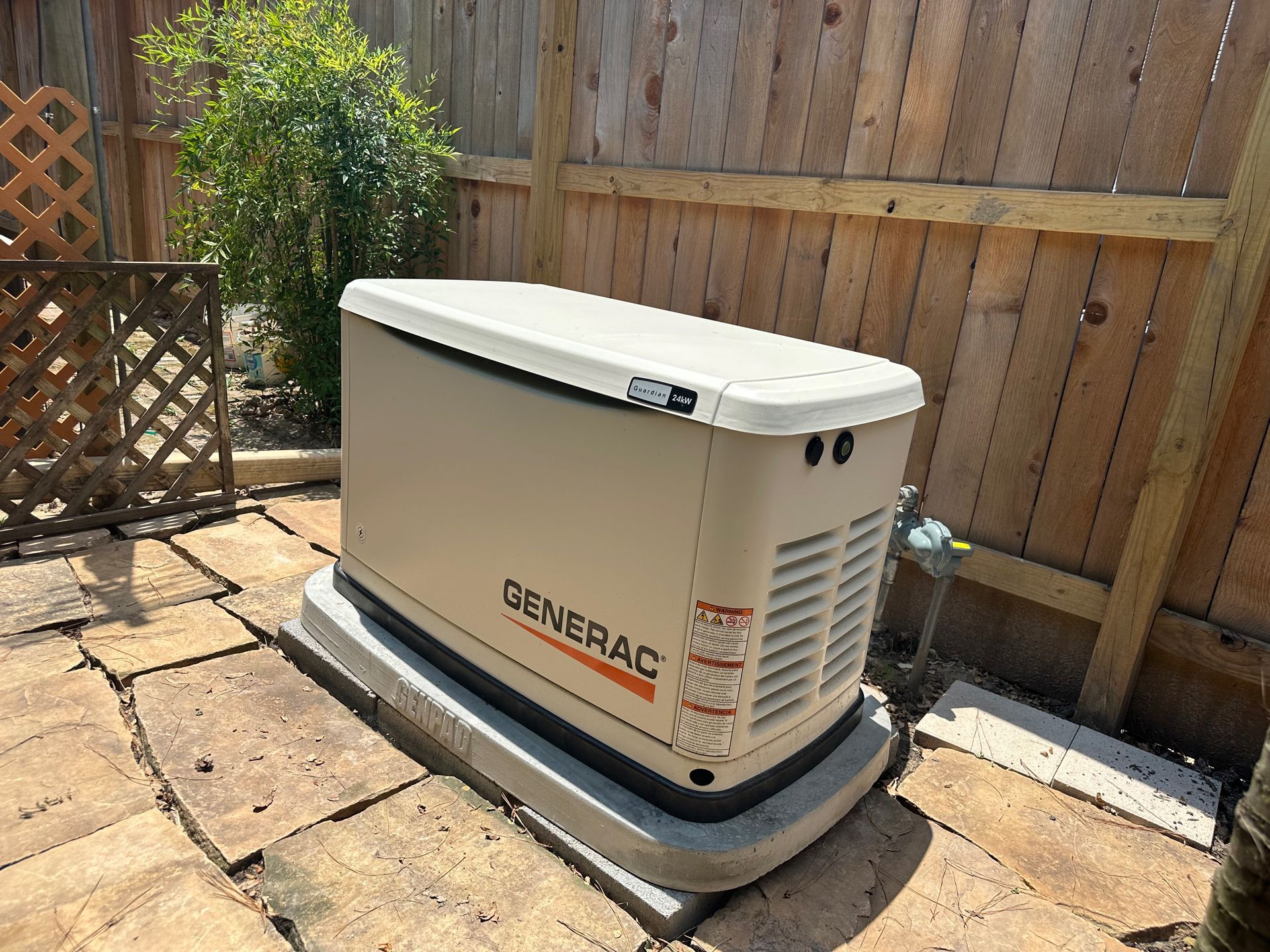 Tan Generac home generator on a concrete pad, surrounded by a wooden fence and paving stones.