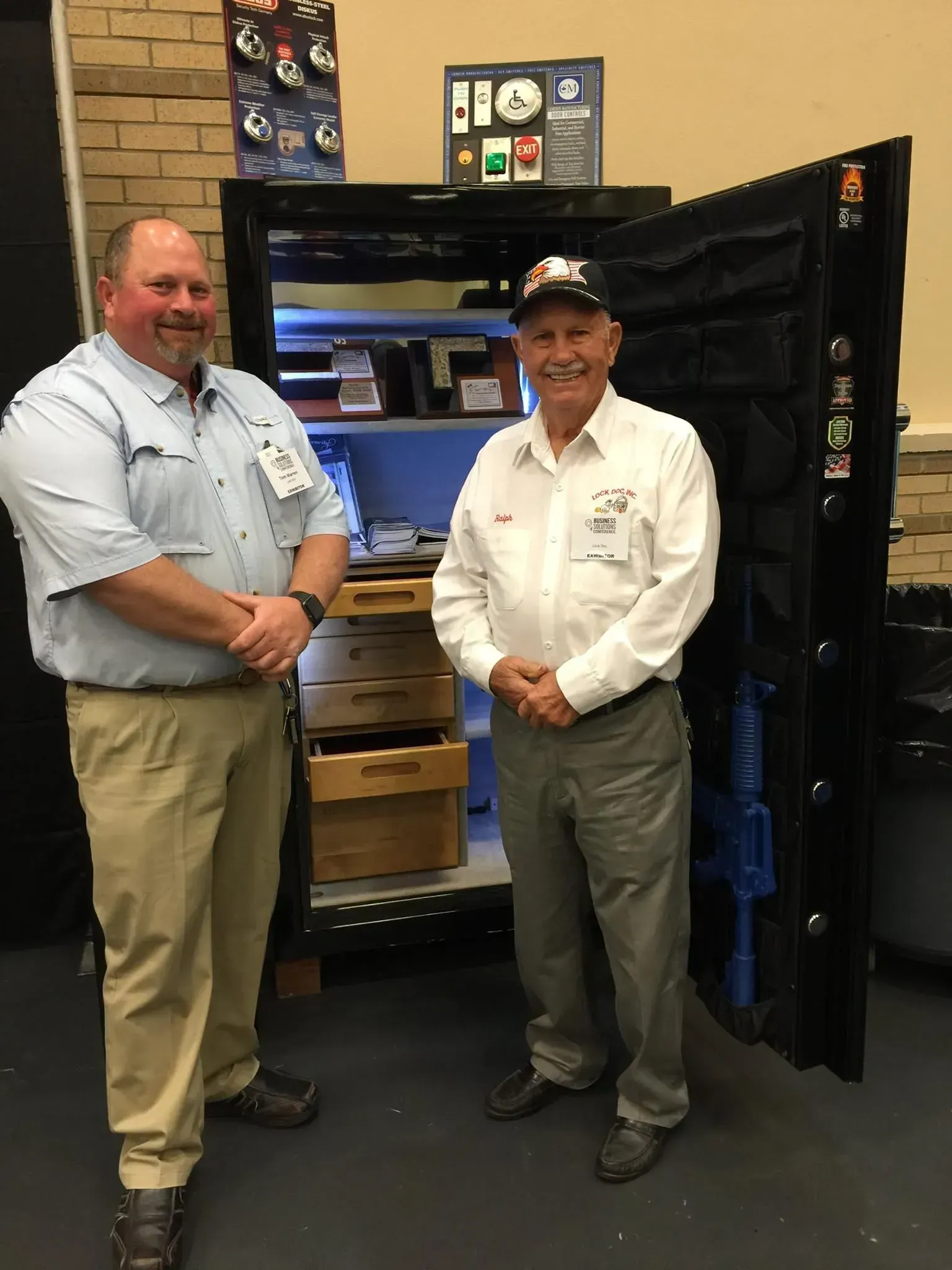 Two smiling men stand in front of an open, well-lit safe displaying drawers and documents. 