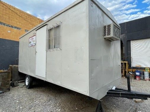Un remolque de oficina portátil está estacionado en un lote de grava frente a un edificio.