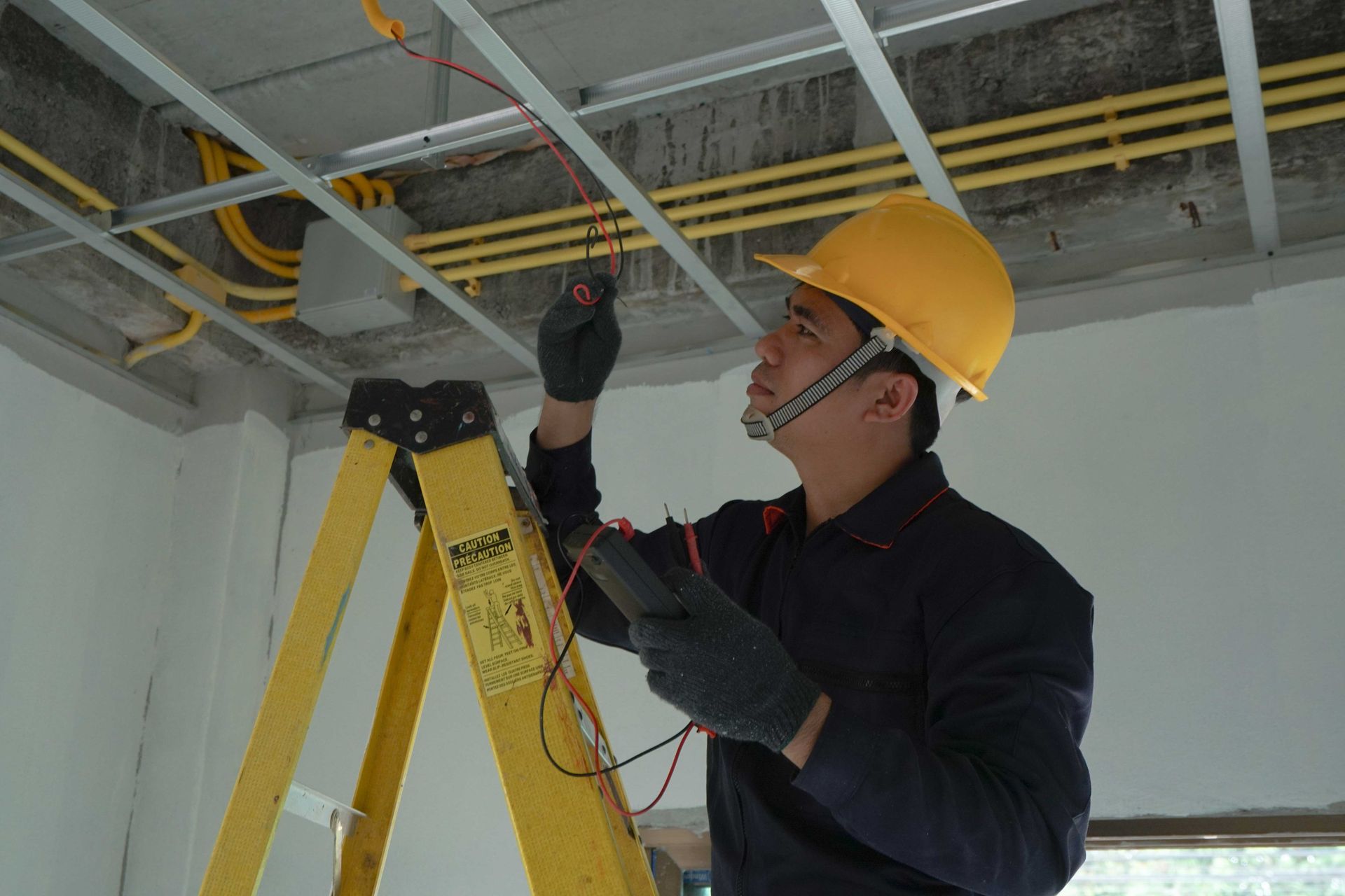 Worker in hard hat inspecting ceiling wiring beside a yellow ladder in an unfinished room