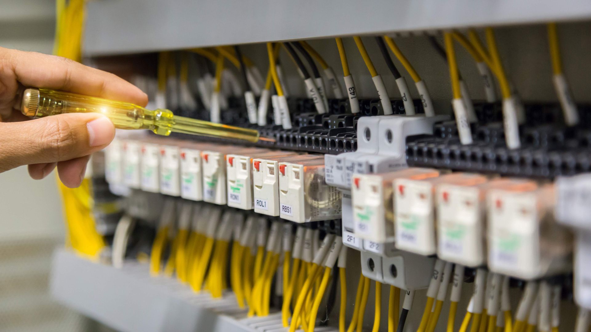 Hand holding a screwdriver near an electrical panel with rows of switches and yellow wires
