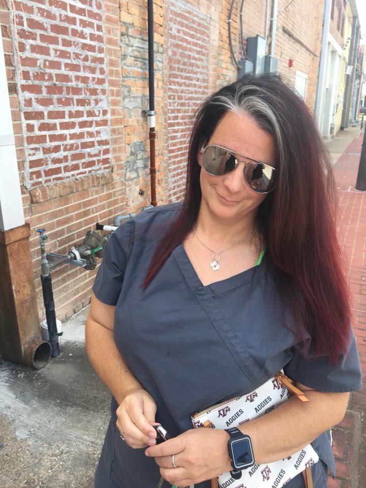 A woman wearing scrubs and sunglasses is standing in front of a brick building.