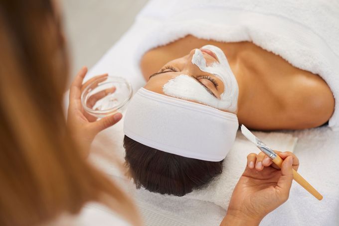 Beautician covering woman facial skin with moisturizing mask during skincare procedure.