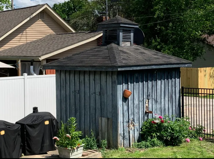 A blue shed with a black roof is in the backyard of a house.