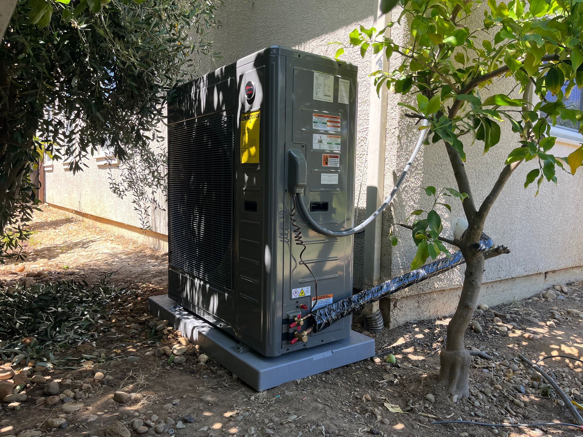Outdoor HVAC unit on a concrete pad beside a stucco wall, partly shaded by trees.