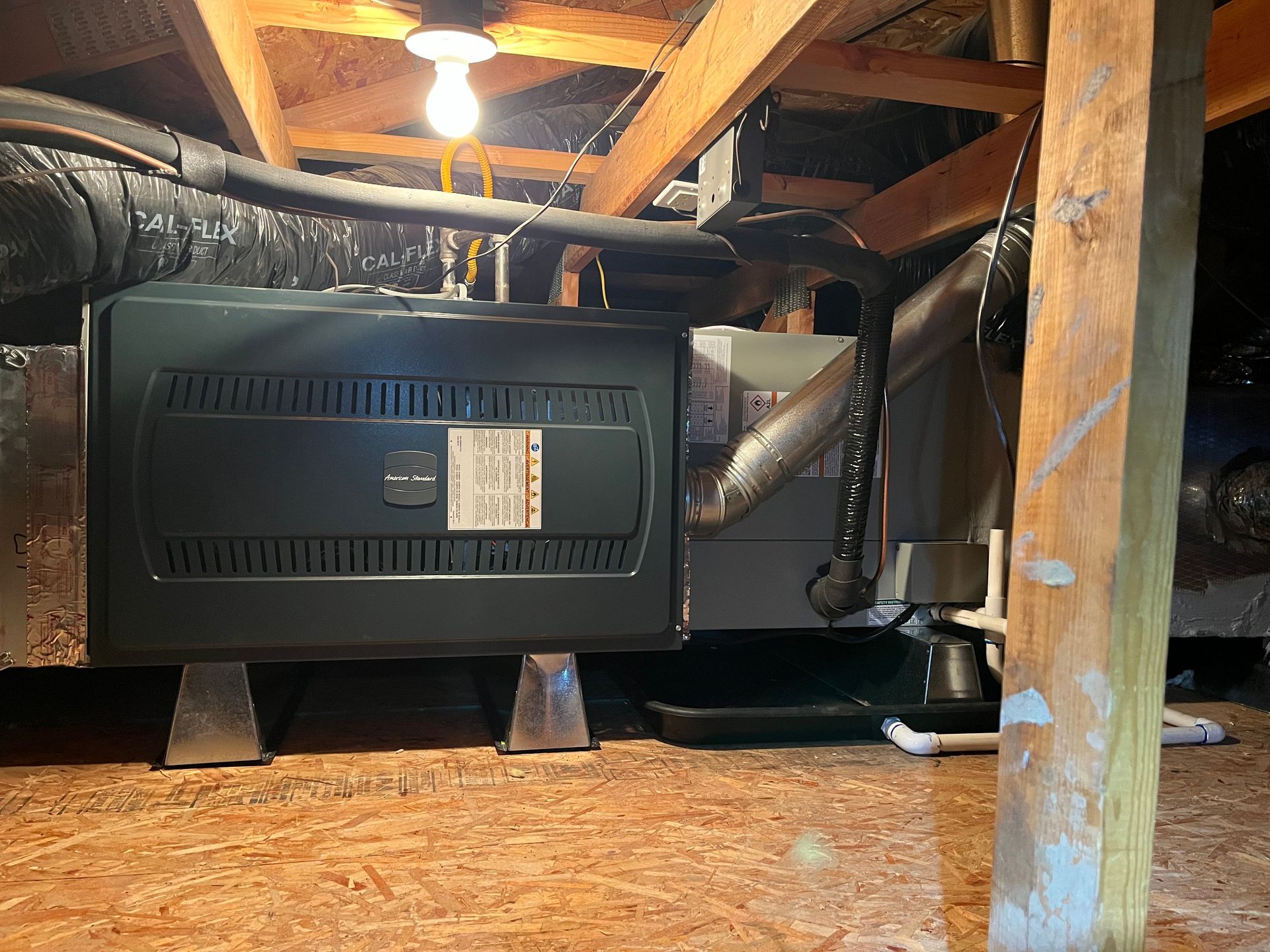 Black furnace under a house with exposed beams, ducts, and a bare light bulb in the crawlspace.