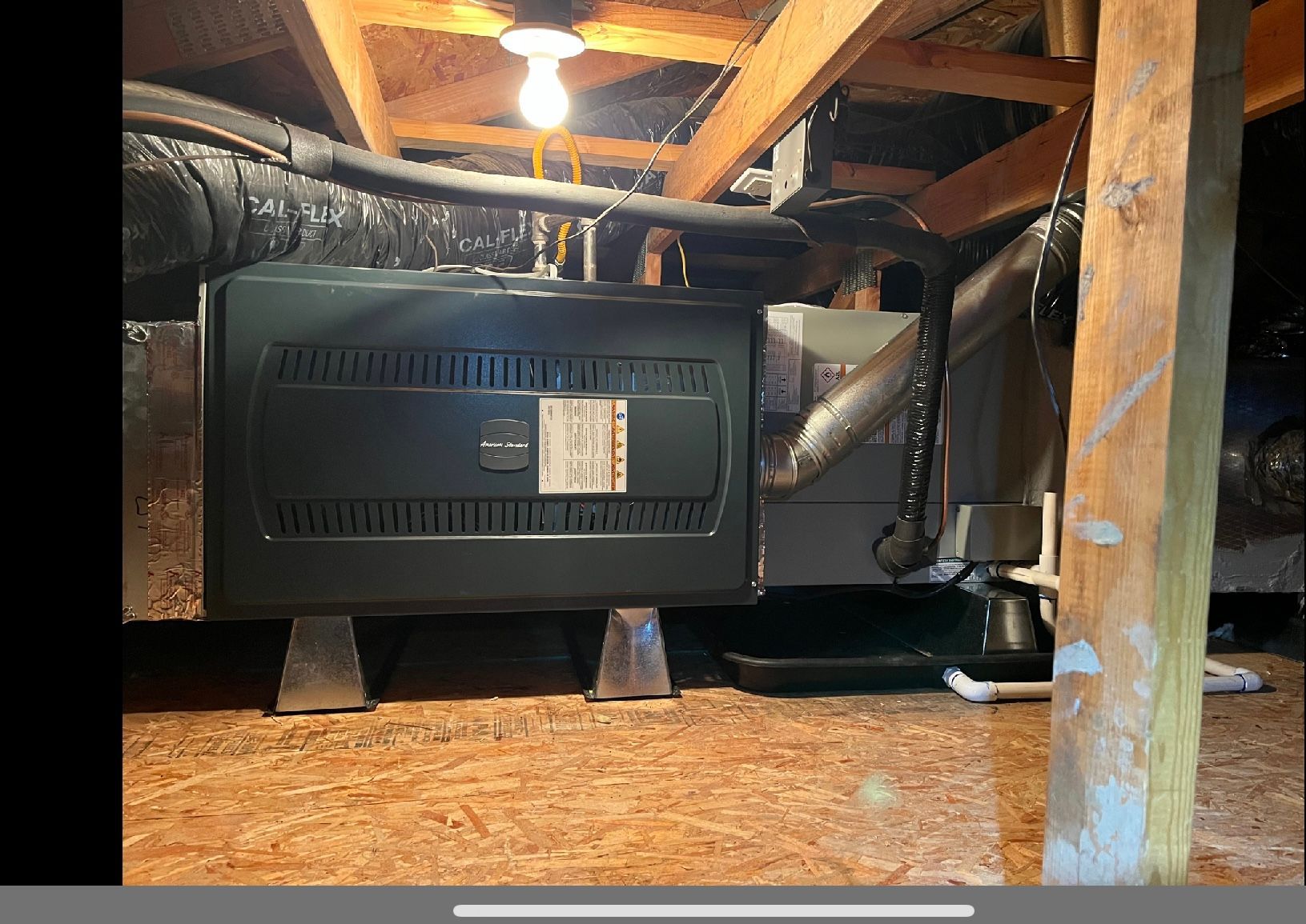 Black furnace unit in a crawl space, with ducts, pipes, and wooden beams overhead.