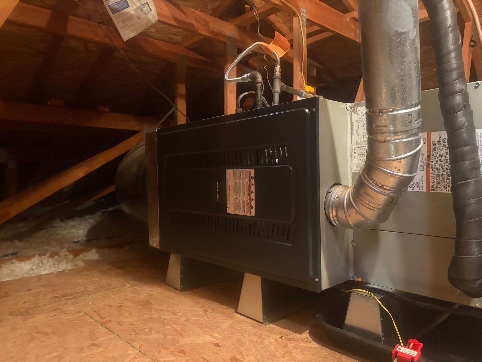 Gas furnace in an attic with silver ducting and exposed wooden rafters.