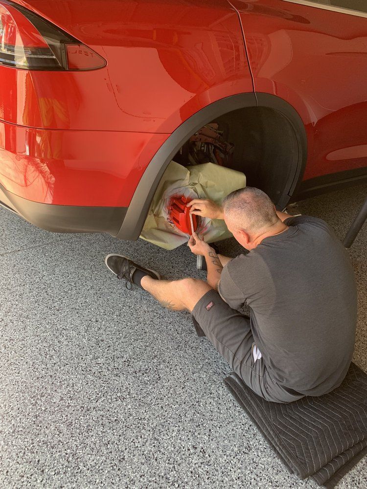 A man is sitting on a mat in front of a red car.