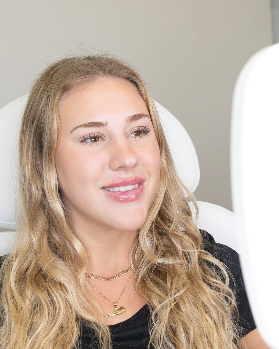 Woman with blonde hair smiling, looking at her reflection in a mirror; medical setting.