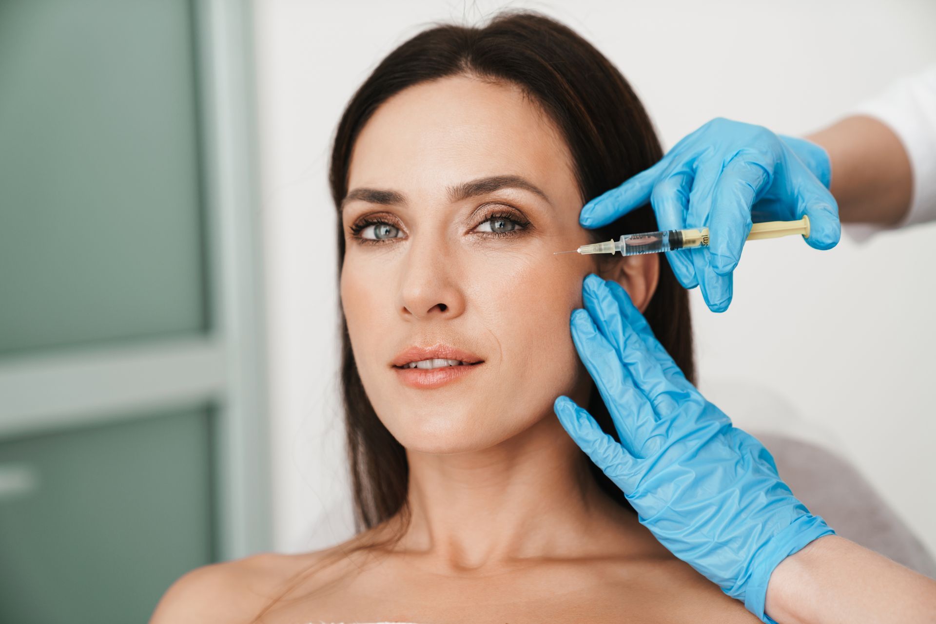 Woman receiving a facial injection in a medical setting, with blue gloved hands holding the syringe near her cheek.