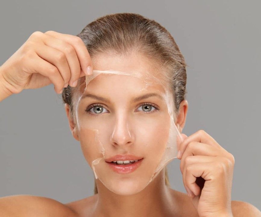 Woman peeling a facial mask from her face, with neutral expression, against a gray background.