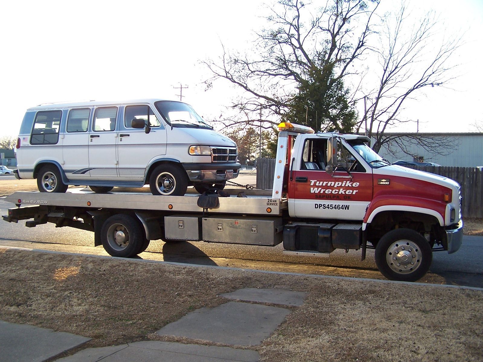 White van on a tow truck. The truck is red and white, parked on a sidewalk next to a road, setting outside during the day.