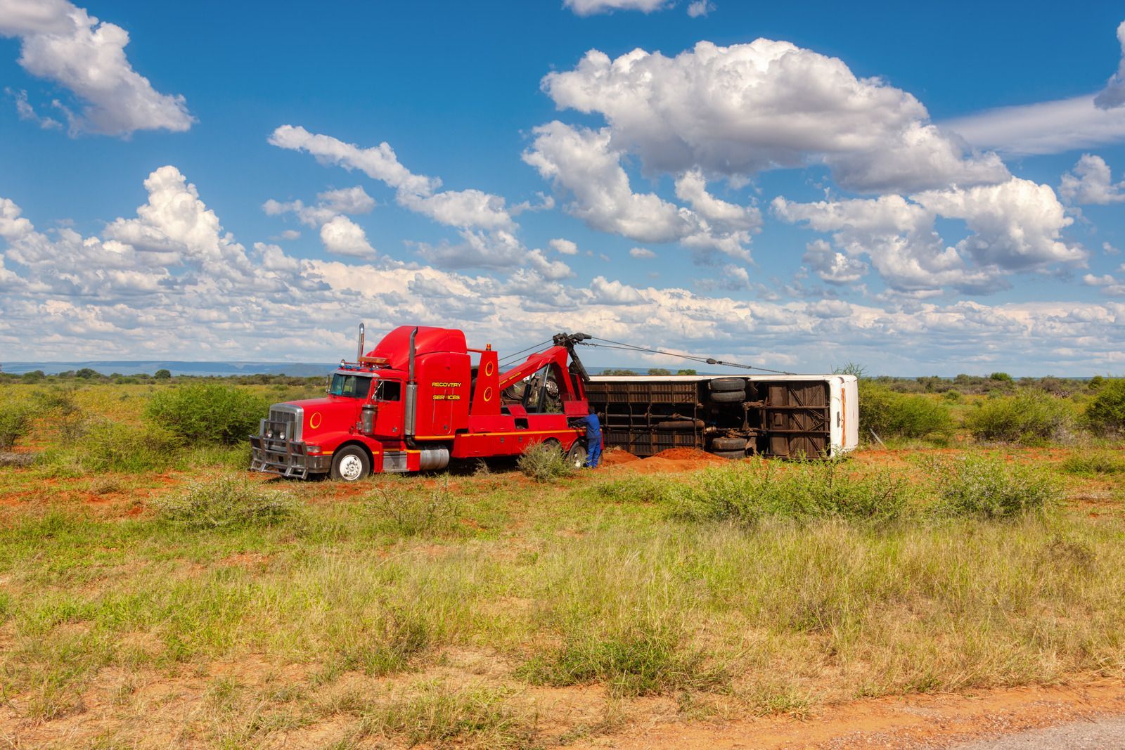 Red tow truck upright, trailer overturned in field under a blue sky with clouds.