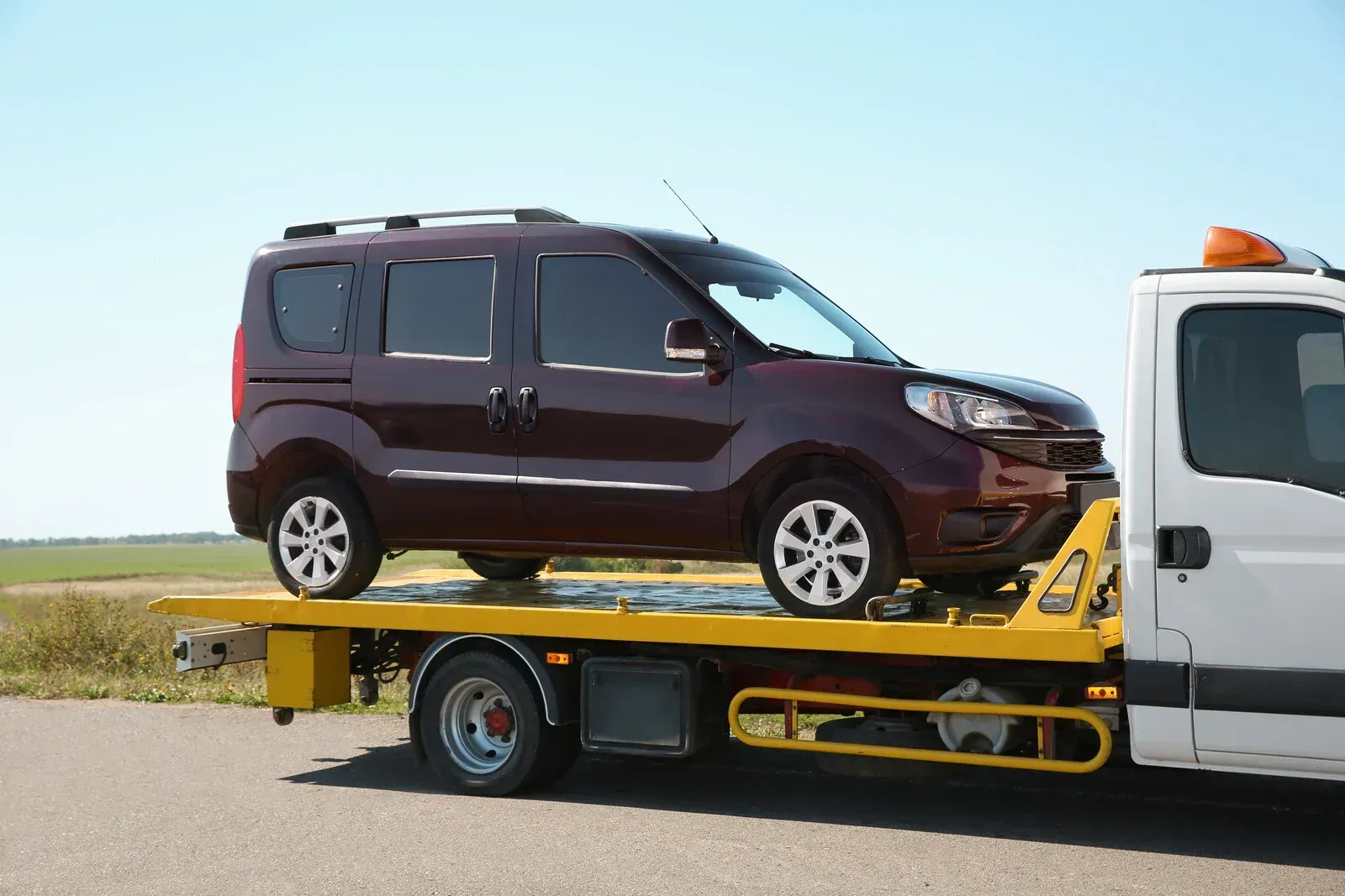 Dark red minivan on a yellow flatbed tow truck. Sunny outdoor setting.