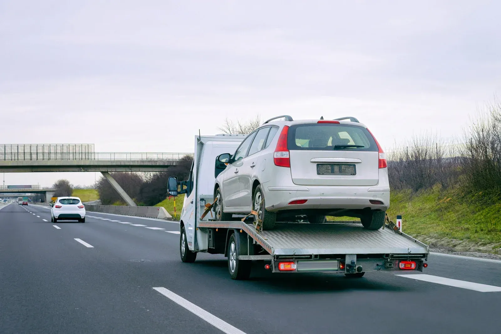 Tow truck carrying a silver station wagon on a highway under cloudy skies.